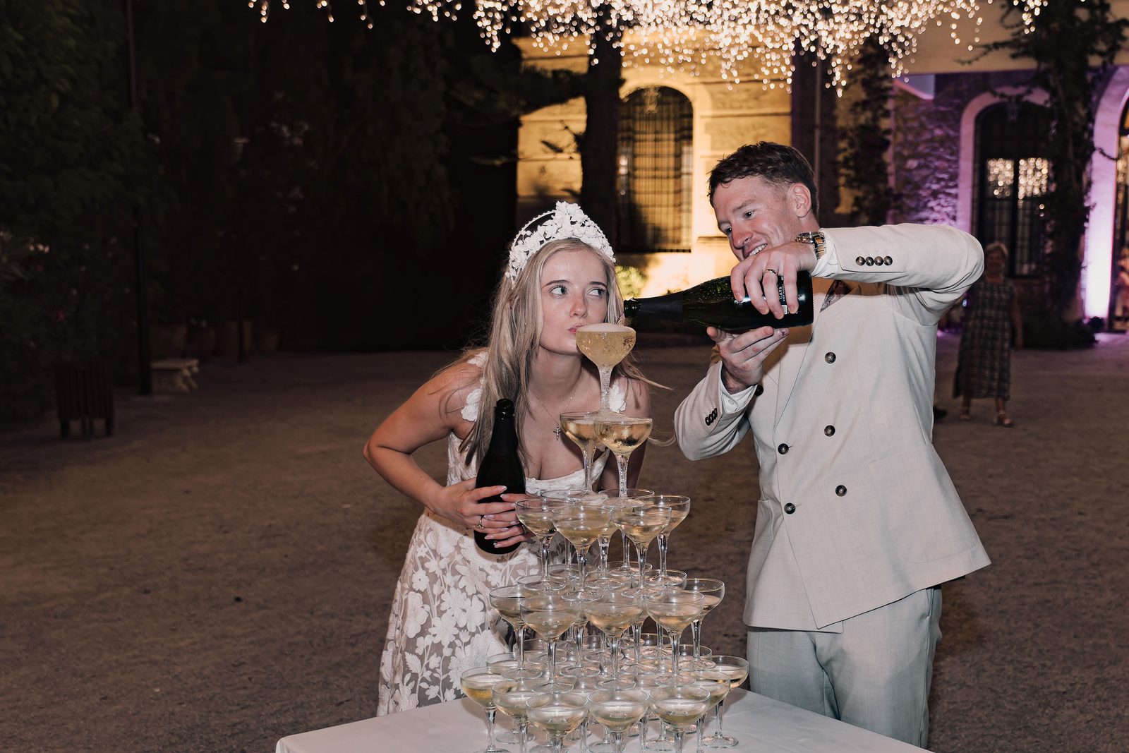 A bride in a tiara drinks from a champagne flute as a groom pours from a bottle above a tower of glasses