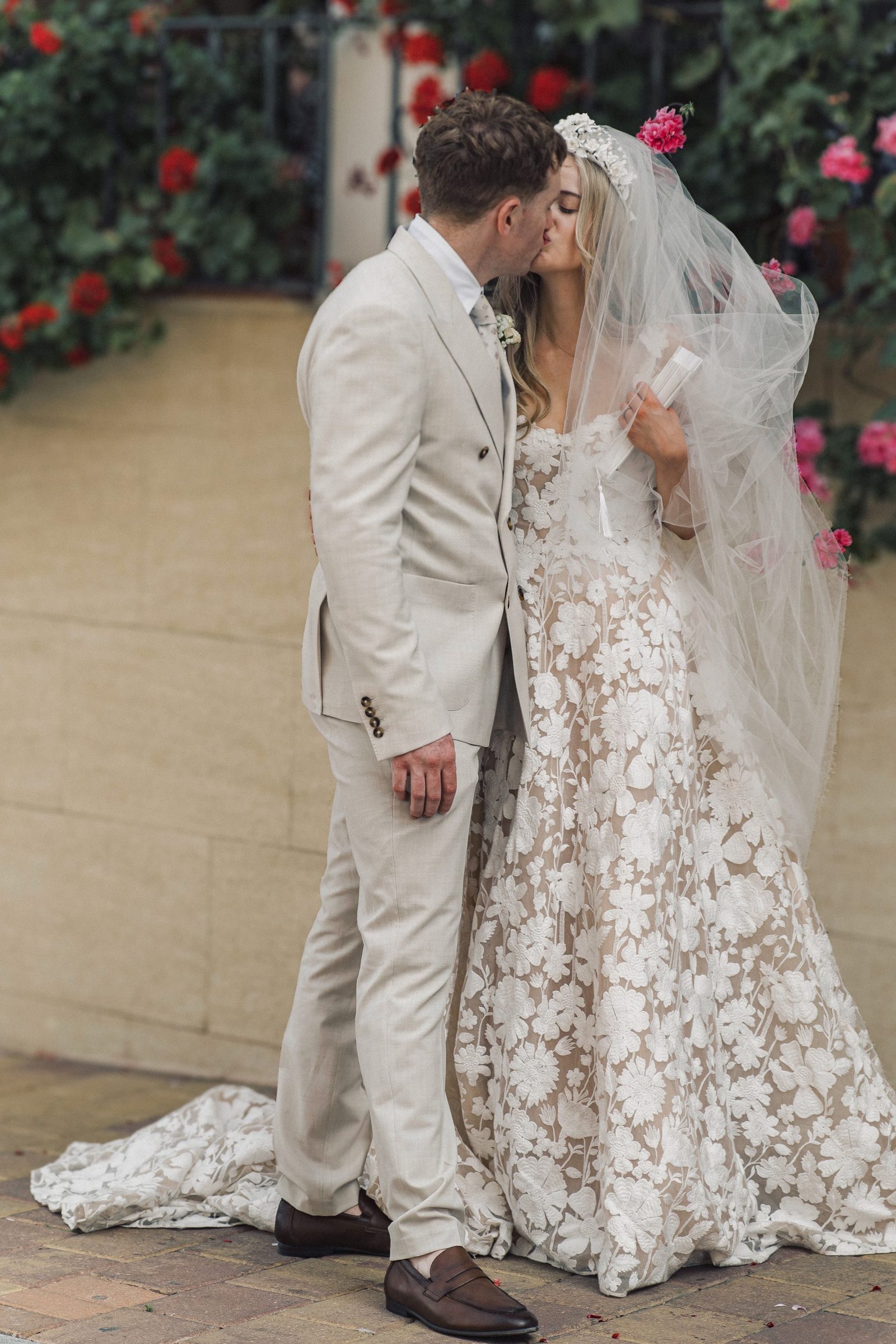 A bride and groom kiss outdoors beside red flowers